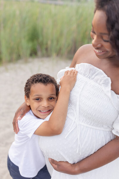 A pregnant mother stands on the beach with her young child, both smiling and enjoying the moment. The golden light reflects off the water, creating a peaceful scene. Captured by a Fairfield photographer, this beautiful maternity moment is set against the serene beach backdrop