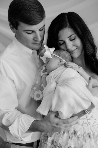 Black and white newborn photo with baby girl laying on mom and dad standing behind mom smiling at baby.