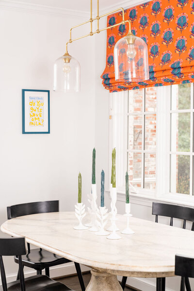 Dining room with light wood table, black dining chairs, orange and blue blinds and a gold light fixture