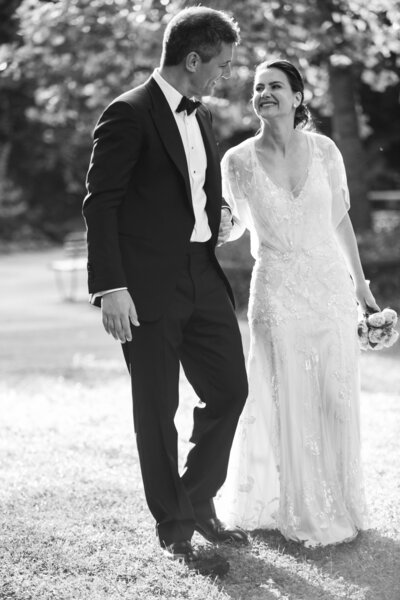Elegant black and white wedding portrait — bride and groom walking outdoors, laughing together in sunlight