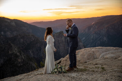 "A groom wipes away tears while reading his vows during a Yosemite elopement at Taft Point at sunset. The bride listens with emotion as the golden light illuminates the vast mountain landscape behind them. Captured by Yosemite elopement photographers, this moment reflects the deep connection of an intimate wedding in nature.