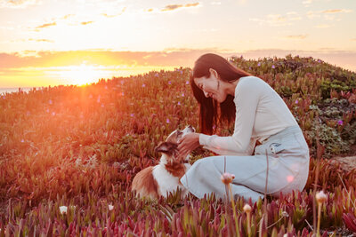 Woman sitting on a field in the sunsent holding dogs head in her hands and smiling