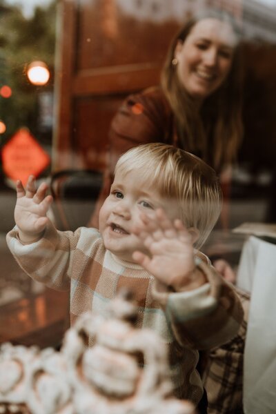 Candid moment of a toddler pressing their hands against a café window, smiling with joy, while a parent looks on warmly in the background.