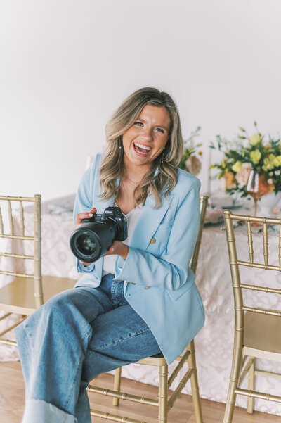 Headshot of Lindsey Kristian sitting in a gold chair wearing a blue blazer and jeans and holding her camera