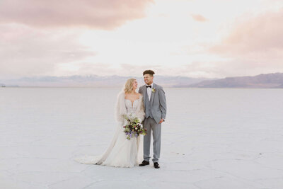 A groom laughs while his bride looks at him while on the Bonneville Salt Flats on their wedding day. 