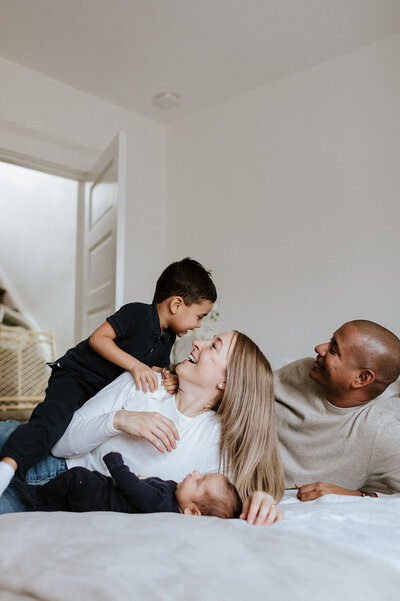 A family of four relaxes on a bed; a woman cradles a baby while a young child sits on her lap, and the man beside them smiles. This heartwarming moment is beautifully captured by a family photographer in Toronto.