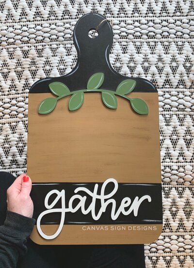 Black wooden cutting board sign with green wreath and white hand lettered "gather"
