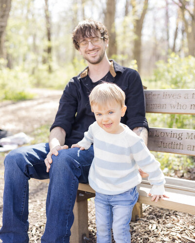 dad and son sitting on a bench with trees in the background, with dad looking down smiling at his son, and son looking at the camera smile