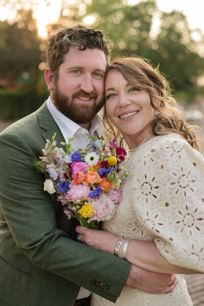 wedding couple posed cheek to cheek with wildflower colorful boquet bride in cream textured dress groom with green suit and bokeh sunset in backgroud