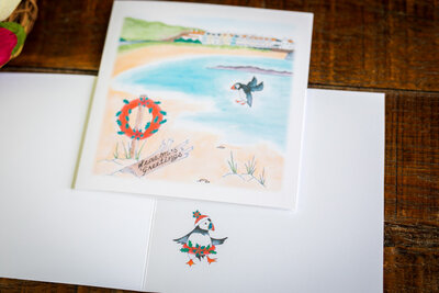 Original watercolour print card featuring puffins over Braye Beach, Alderney