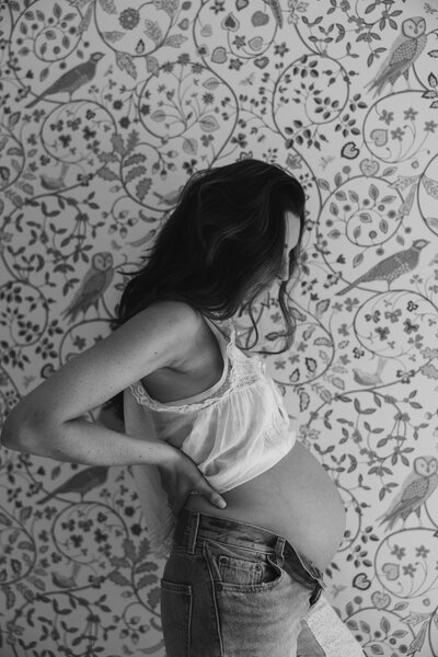 A black and white photo of a pregnant woman standing sideways, gently cradling her belly with one hand and supporting her back with the other. She is in front of a beautifully patterned wallpaper featuring birds and flowers, highlighting the serene and artistic nature of the moment.