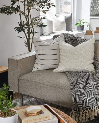 Two decorative off-white pillows and a black and white pattered blanket on top of a modern grey couch in a bright living room filled with plants