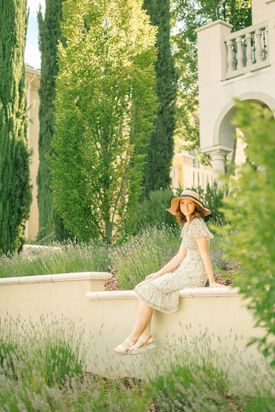 girl in white dress and floppy sun hat seated on  the wall of a lush garden