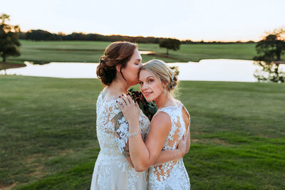 Brides hold each other closely while overlooking pond at wedding venue