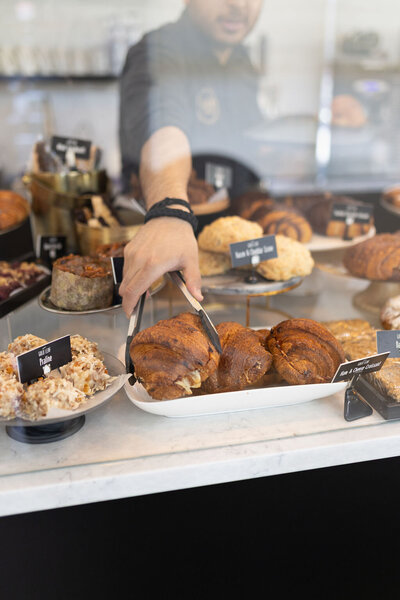 Hand serving a fresh croissant from a bakery display filled with assorted pastries at Gold Leaf Pastries in Kamloops BC.