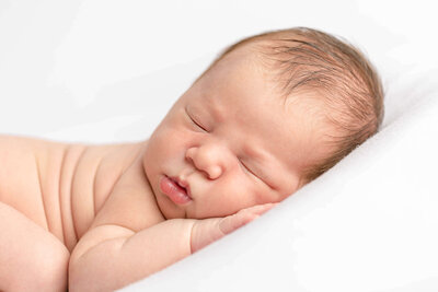 Close-up image of a newborn baby face. Baby is sleeping peacefully on his belly and has one hand up under his cheek. He has a little bit of dark hair on the top of his head.