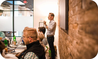 Michael Hyatt standing at the front of a conference room, speaking to a group seated around a table.