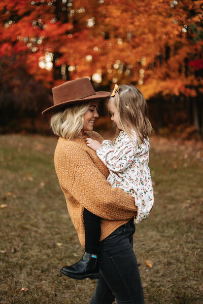 Mother holding daughter facing each other with beautiful fall folliage behind them.
