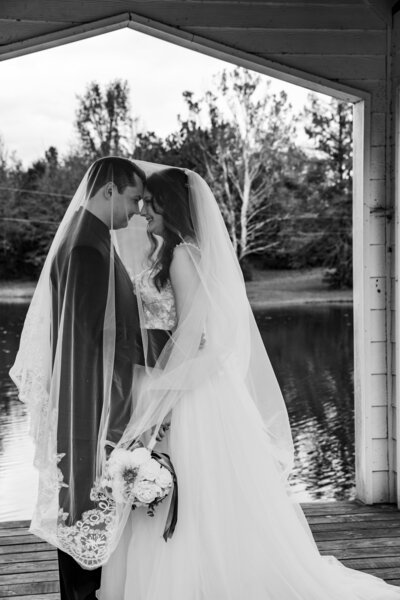 Black and white image of bride and groom standing under a gazebo over a lake, both smiling under her long veil.