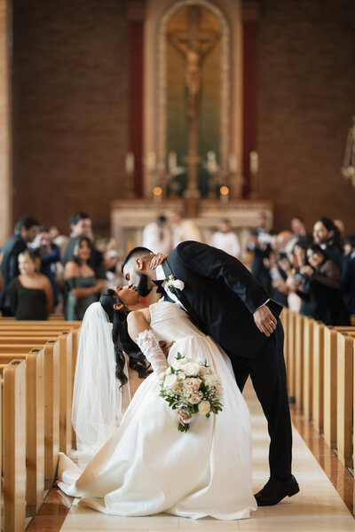 Couple kissing on their wedding day while the wedding party cheers behind and around them.