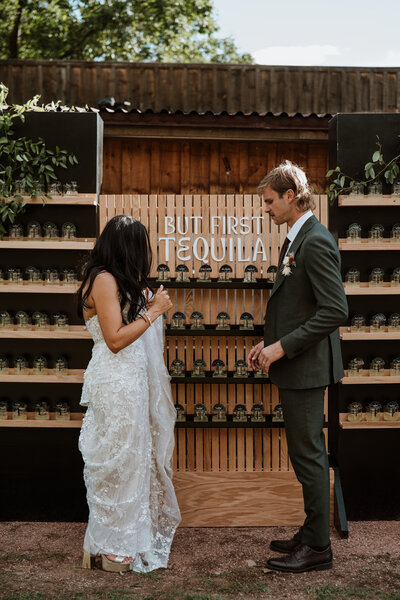 Bride and groom standing at a “But First Tequila” shot wall at their Breckenridge wedding.