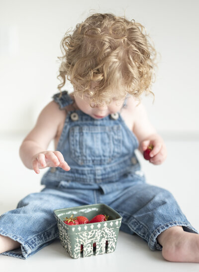 Toddler in denim overalls sitting indoors, focused on eating strawberries.