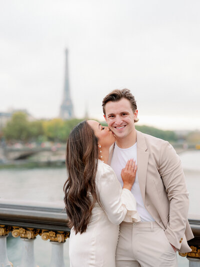 a man in a tan suit being kissed on his cheek by a woman in a white dress standing on the [ont alexandre bridge with the eiffel tower behind them