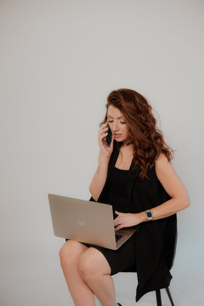 Amy the owner of a bridal hair stylist business sitting on a stool in a studio in front of a white background wearing an peach or off white blazer