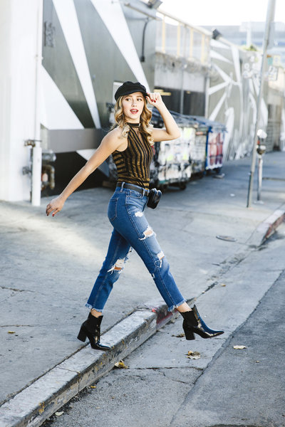 High-energy urban senior portrait of a teen girl mid-step in ripped jeans, black boots, and a striped top. Captured with an edgy downtown LA street-art backdrop for an editorial vibe.