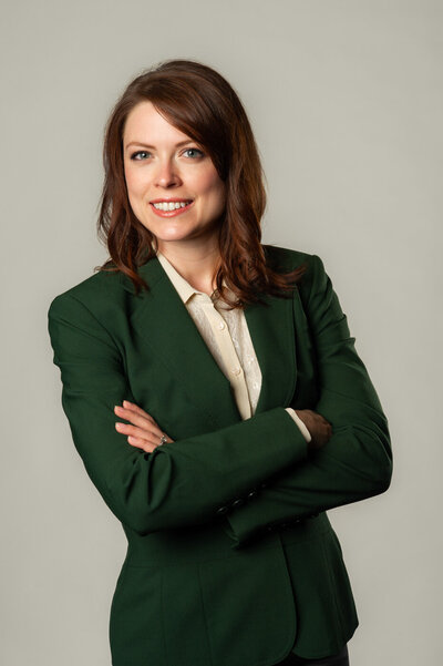 Studio headshot of a beautiful business woman with reddish hair and dark green blazer smiling with arms crossed.