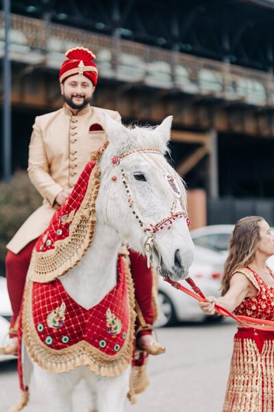 A groom in traditional attire riding a decorated white horse with a person leading the horse by its reins.