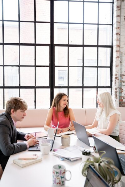 Pierce PR team members collaborating at a white table with laptops, coffee mugs, and notebooks in a bright industrial-style office with large grid windows.