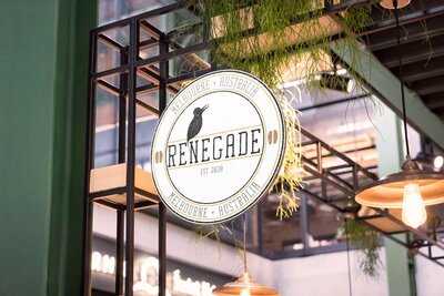 A circular sign hanging from a black metal frame with plants hanging above. The sign reads renegade established 2020 Melbourne Australia. On top of the word renegade a bird sits with two coffee beans on either side.