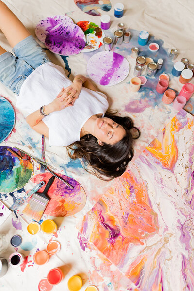 Creative Director and artist Courtney Claire Beglin lays on the ground surrounded by her paintings with her eyes closed, showing her expansive imagination