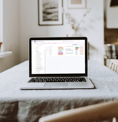 An open laptop on a linen-covered table in a bright room displays the Get Shit Done Hub on the screen: a database-style task tracker containing rows with colour-coded labels and status fields.