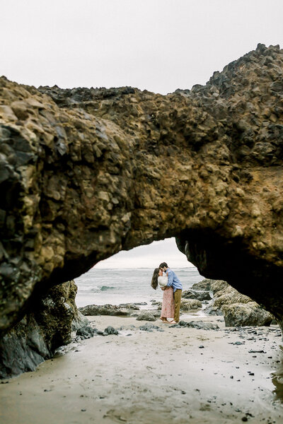 A couple kissing on the beach, framed by rocks, representing the shooting style of lifestyle photographer Among the Oaks. 
