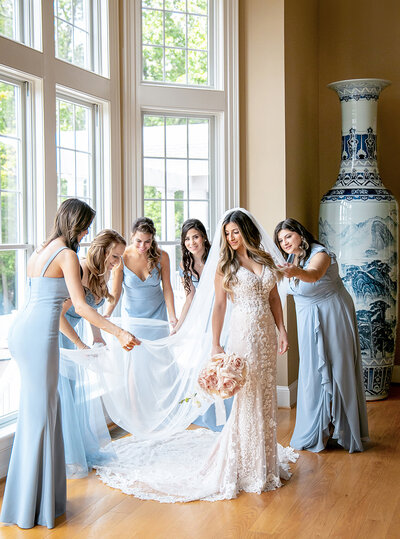 Bride in a lace gown stands by tall windows, smiling as five bridesmaids in light blue dresses adjust her veil. Large porcelain vase in the background. Bright natural light floods the room, highlighting the elegant scene.