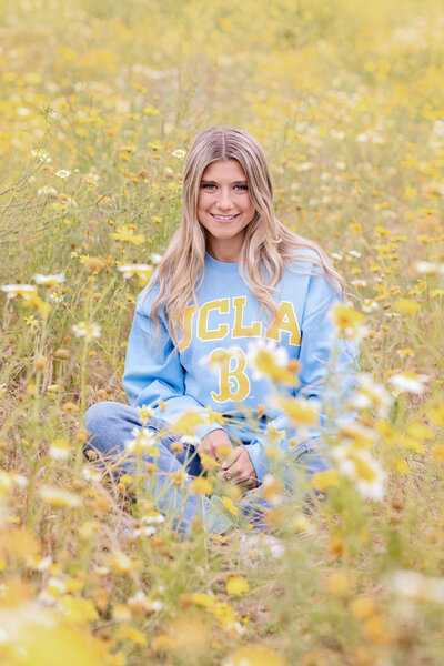 A blonde young woman wearing a blue UCLA sweatshirt giving the "fours up" sign while standing in a field of yellow flowers