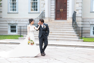 A romantic wedding photo of a groom kissing his bride's hand outside a grand estate. The bride, in a flowing white gown, and the groom, in a classic black suit, share a tender moment on the steps, capturing timeless love and elegance.