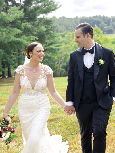 A bride in a lace wedding dress and a groom in a tuxedo smile at each other while walking hand in hand outdoors, with greenery and trees in the background.