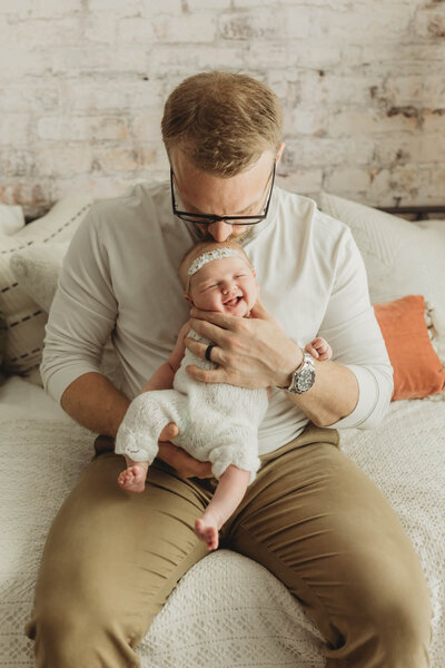 baby girl in a white romper smiling while dad holds her and kisses the top of her head