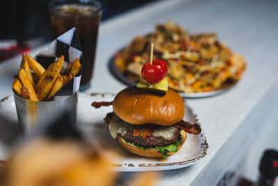 Delicious bacon cheeseburger topped with a pickle and cherry pepper, served with crispy golden fries and a refreshing soda, with a plate of loaded nachos in the background.