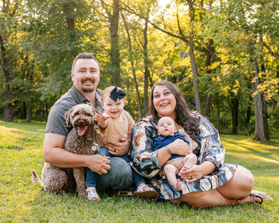 Family with toddler, infant and their dog sitting on the grass for a family portrait.