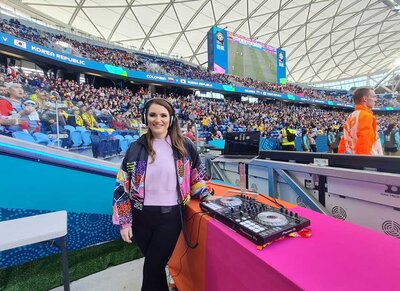 Female DJ with headset and Pioneer deck standing beside the pitch at a packed FIFA Women’s World Cup stadium