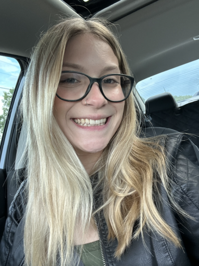 Blonde with nose ring and black rimmed glasses sits in car and smiles at camera