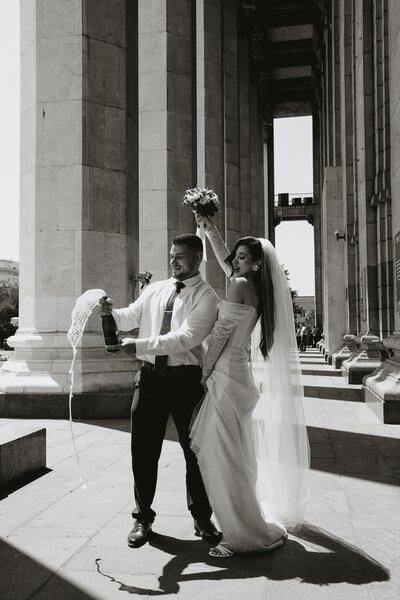 A groom popping a bottle of champagne with a bride celebrating next to him.