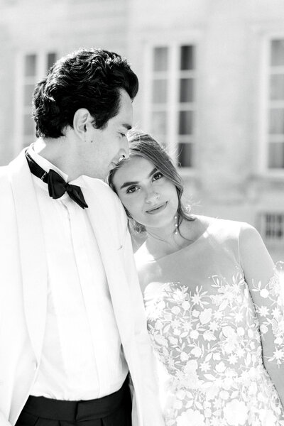 bride leaning on her groom's shoulder in front of a french chateau groom wearing a white dinner jacket and bride in a lace gown