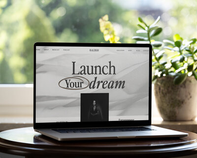 A silver laptop sitting on a desk with the Raleigh Showit Template by Lily on the screen.  Table and laptop are in front of a window and next to a potted plant.