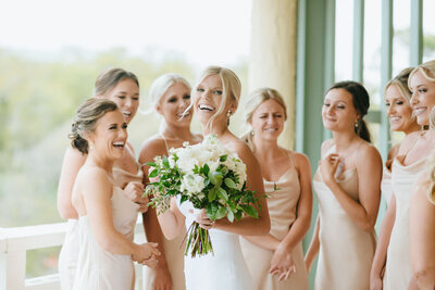 A radiant bride holding a lush white and green bouquet shares a joyful moment with her bridesmaids at The Westin Hotel Savannah. Her smile captures the excitement of the wedding day, surrounded by her closest friends in elegant champagne-colored dresses. Photographed by Amia Marcell Photography.