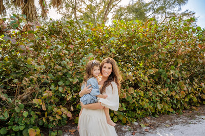 Mother and daughter standing in a beach pathway with greenery behind them. Mom is holding the daughter on her hip and they are both smiling at the camera.